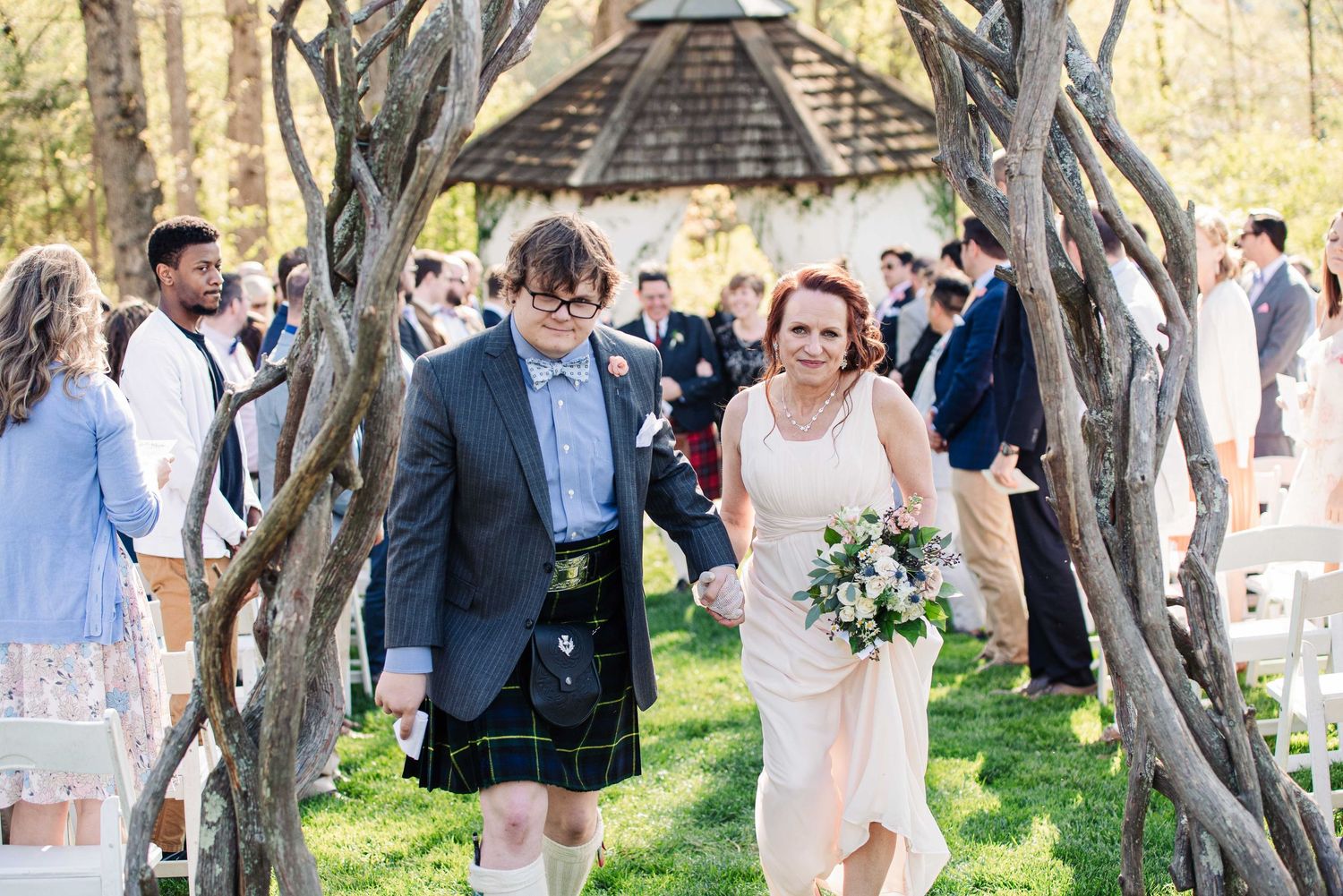 Couple walks hand-in-hand under rustic wooden arch at outdoor wedding ceremony with guests in background.