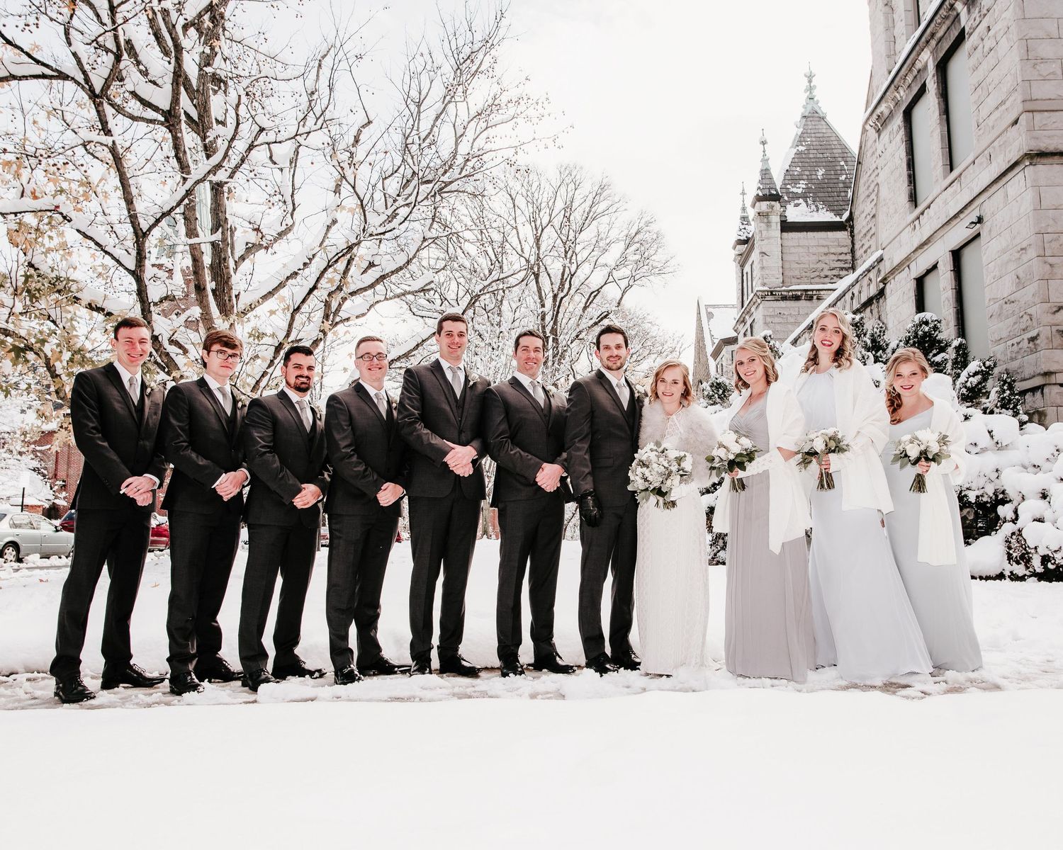Wedding party posing in snow, groomsmen in dark suits and bridesmaids in gray gowns with bride in white dress.