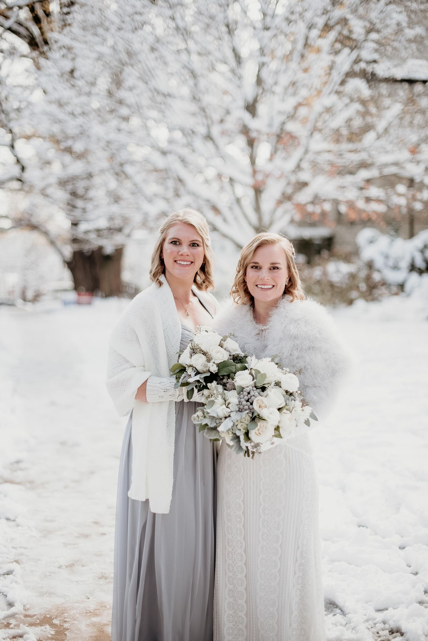 Two brides in white and gray gowns with fur wraps hold bouquets outdoors in a snowy winter wedding setting.