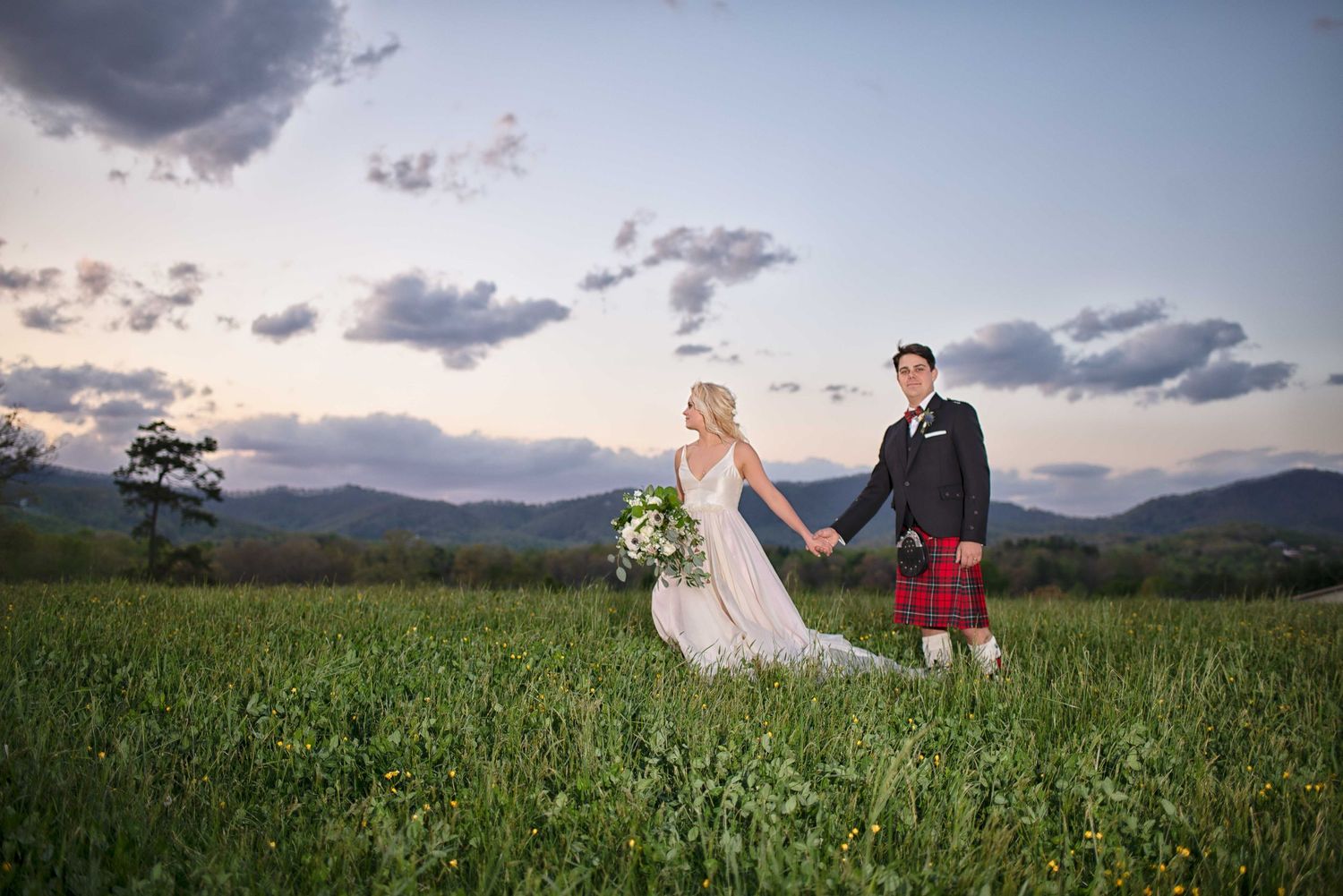 Bride and groom holding hands in green field at sunset, groom wearing red tartan kilt, mountains in background.