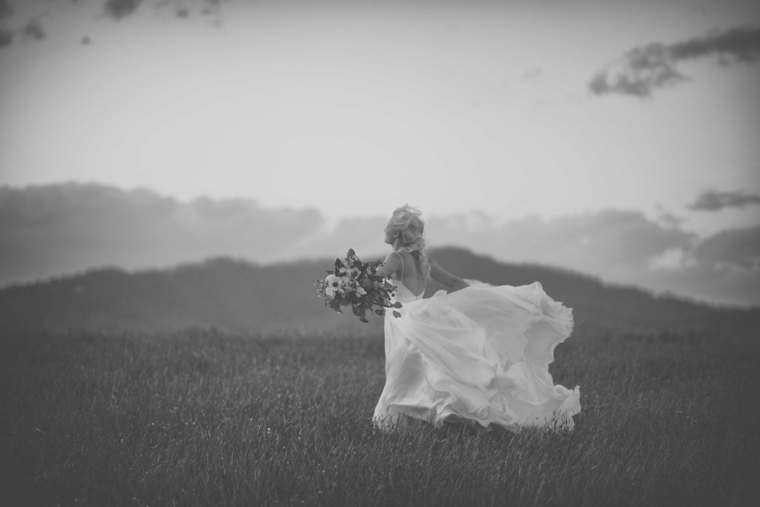Black and white photo of a bride in a flowing white dress holding flowers in a scenic field with mountains.