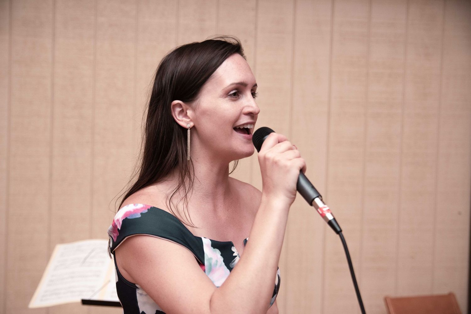 Young woman with dark hair singing into a microphone at an indoor performance event.