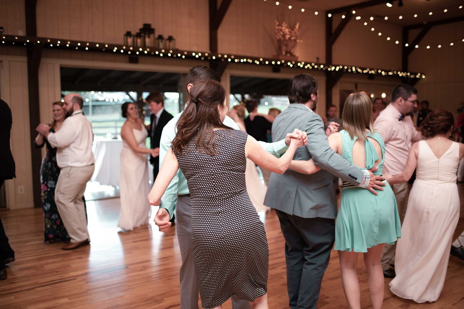 Guests dancing together at a rustic barn wedding reception with string lights and hardwood floors.