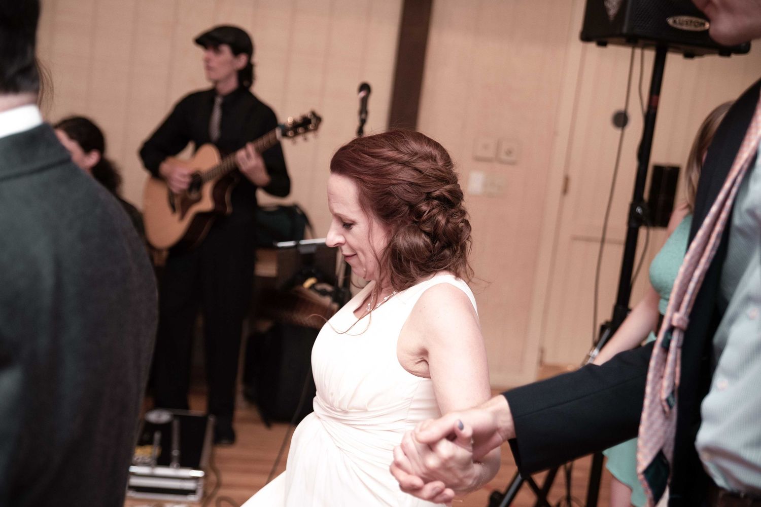 Bride in white dress dancing with partner at wedding reception while guitarist performs on stage.