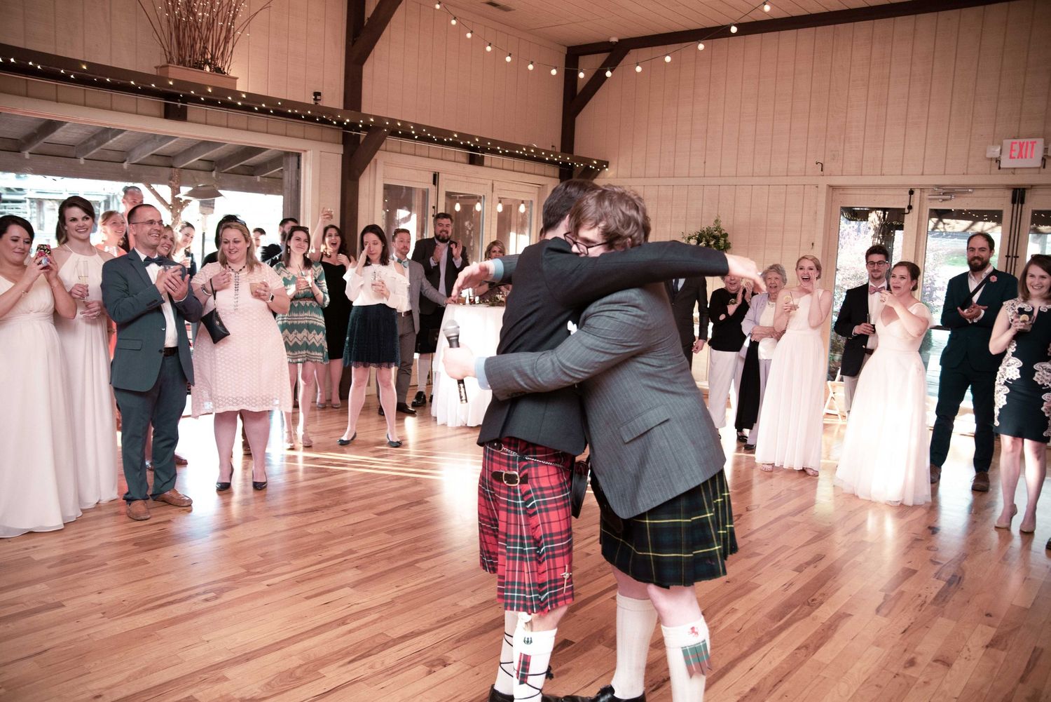 Couple shares first dance in rustic wedding venue, groom wearing traditional Scottish kilt, surrounded by applauding guests.