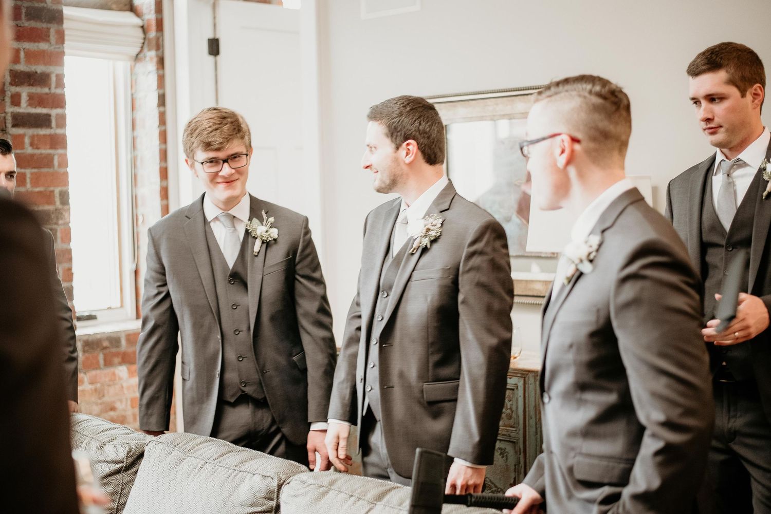 Groomsmen in gray suits with boutonnieres laughing and conversing before a wedding ceremony indoors.