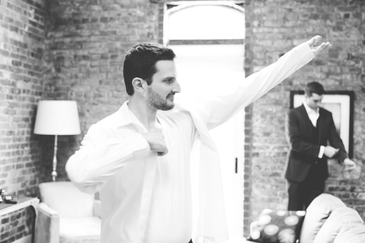 Black and white photo of a groom putting on his dress shirt while a groomsman adjusts his suit in the background.