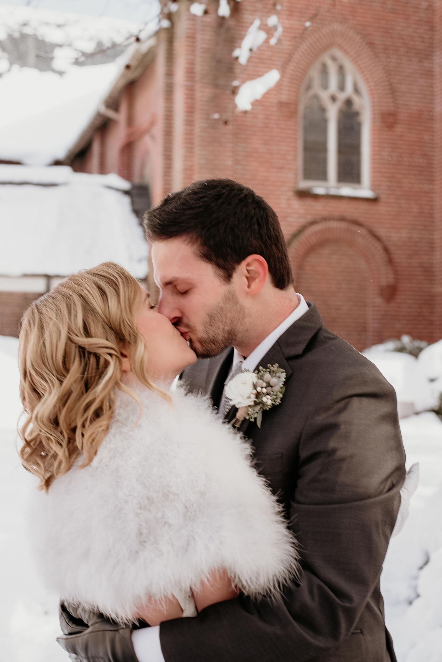 A bride and groom share a romantic kiss outside a snowy brick church in winter wedding attire.
