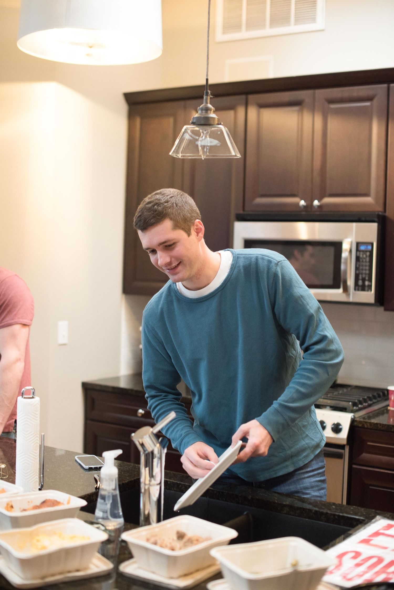 Smiling young man in blue sweater serving food in a modern kitchen with dark wood cabinets.
