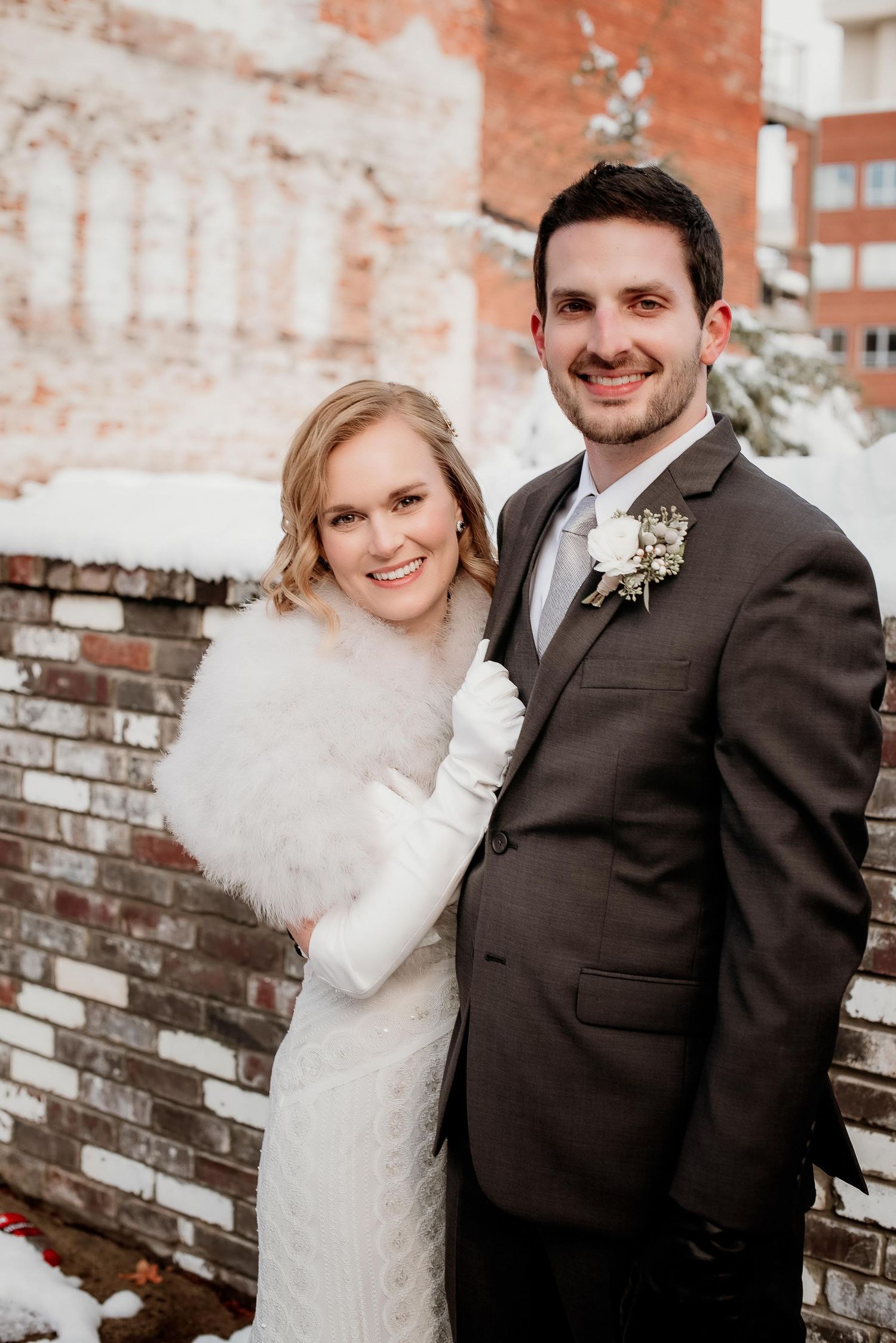 Bride in white lace gown with fur stole and groom in gray suit pose together outdoors in winter snow.