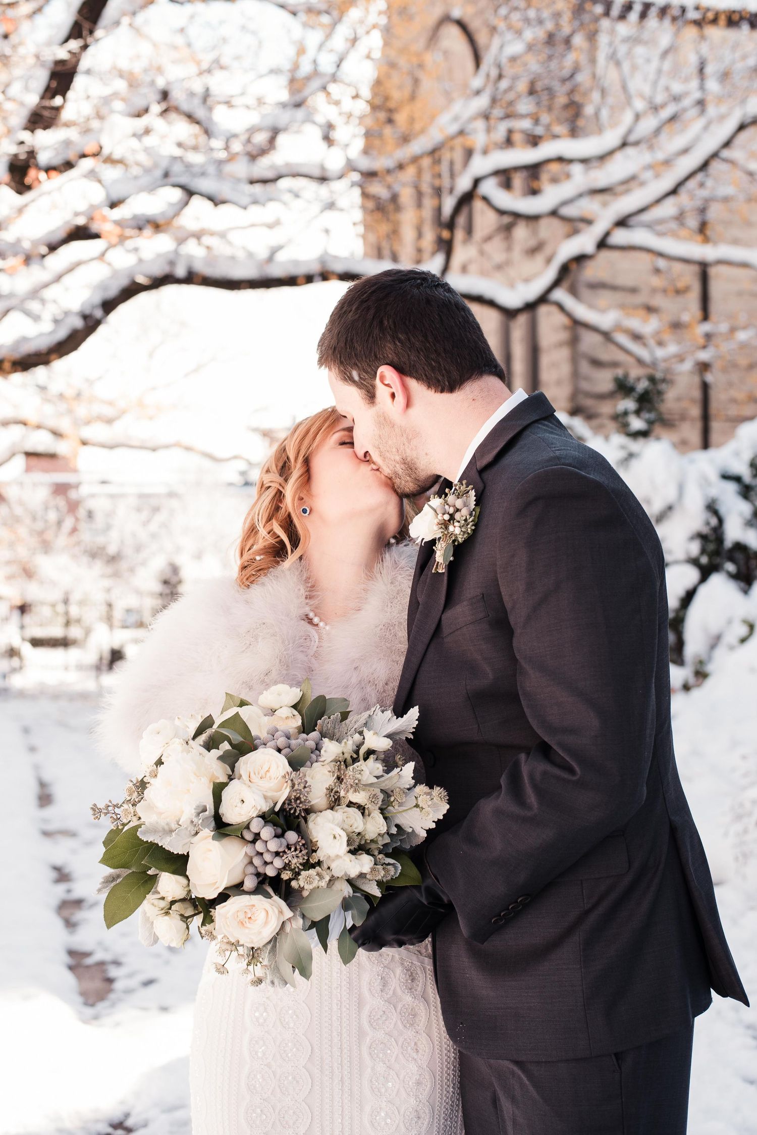 Bride and groom share a kiss in a snowy winter wonderland, bride holding a white floral bouquet.