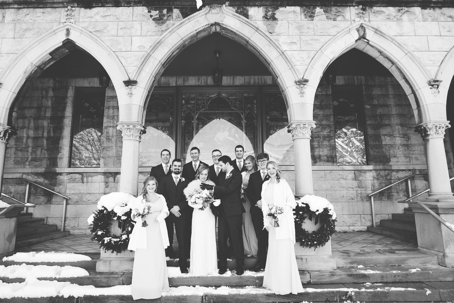 Black and white photo of a wedding party posing on church steps with Gothic arches and snowy surroundings.
