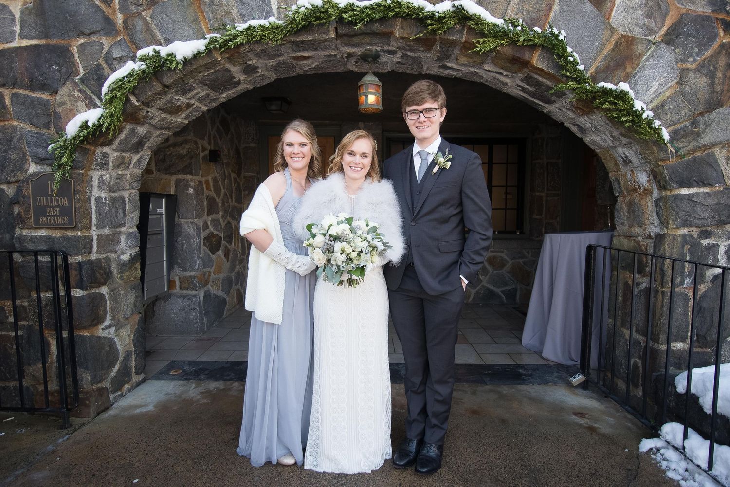 Bride in white gown poses with bridesmaid in gray dress and groomsman under stone arch in winter wedding setting.