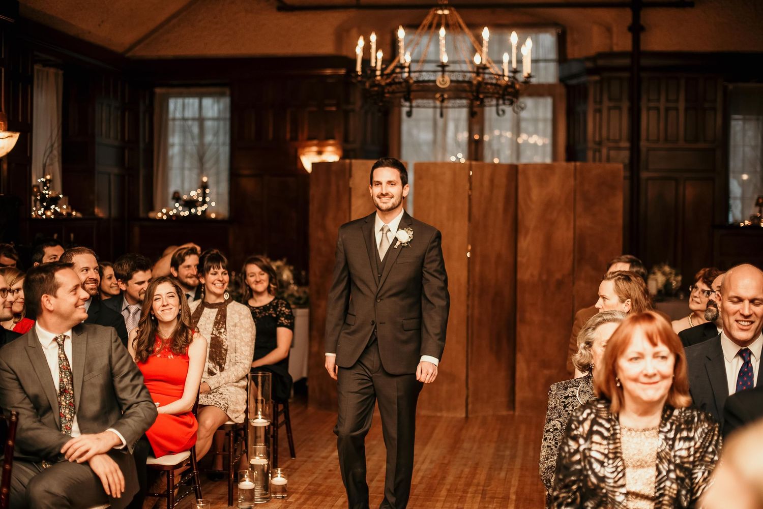 Groom in dark suit smiling at wedding ceremony aisle in elegant wood-paneled venue with chandelier and seated guests.