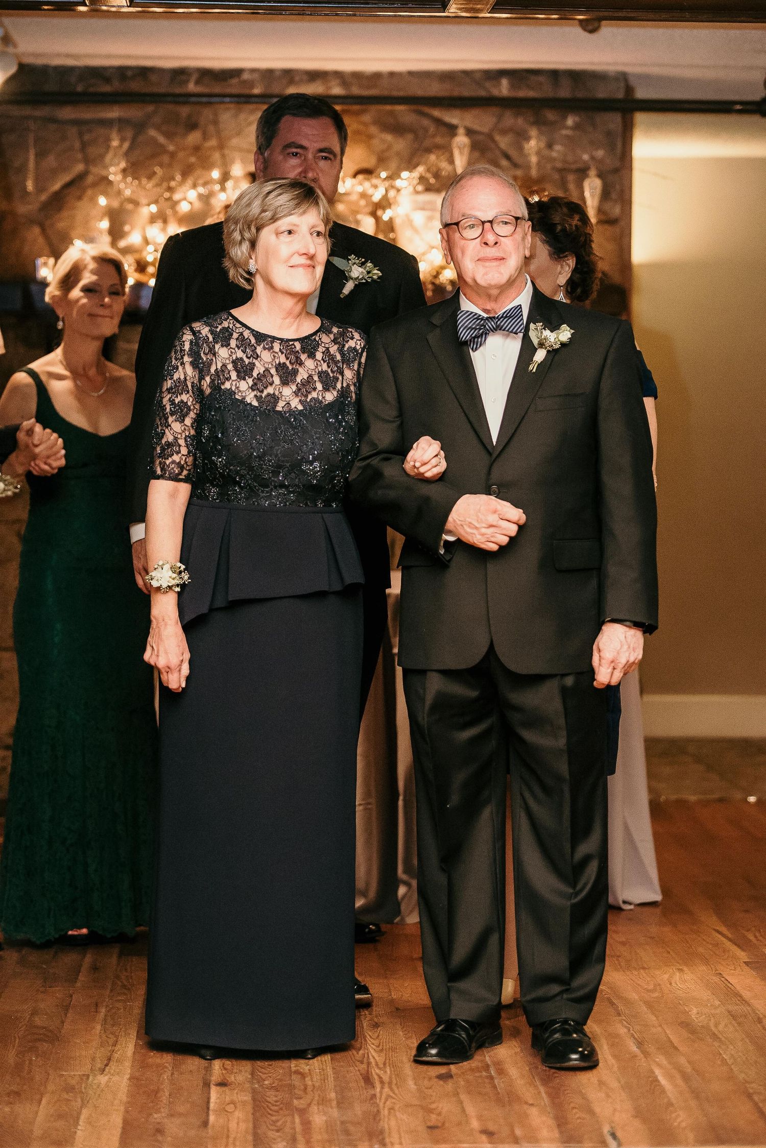 Elegant couple in formal attire at a wedding reception, woman in black lace gown, man in dark suit with bow tie.