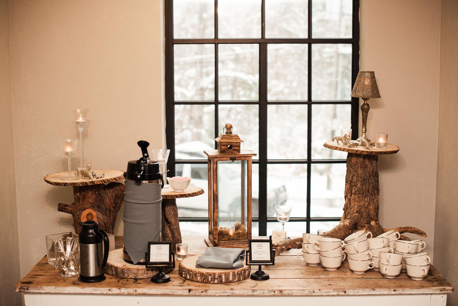 Rustic wedding coffee station with wood stumps, lantern, coffee urns, and white cups on a wooden table.