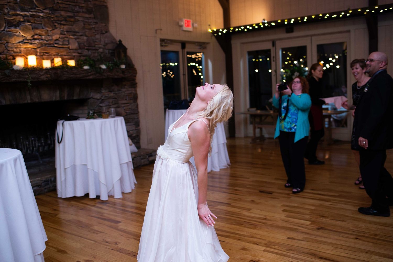 Bride in elegant white gown leans back dramatically on wedding reception dance floor with guests in background.