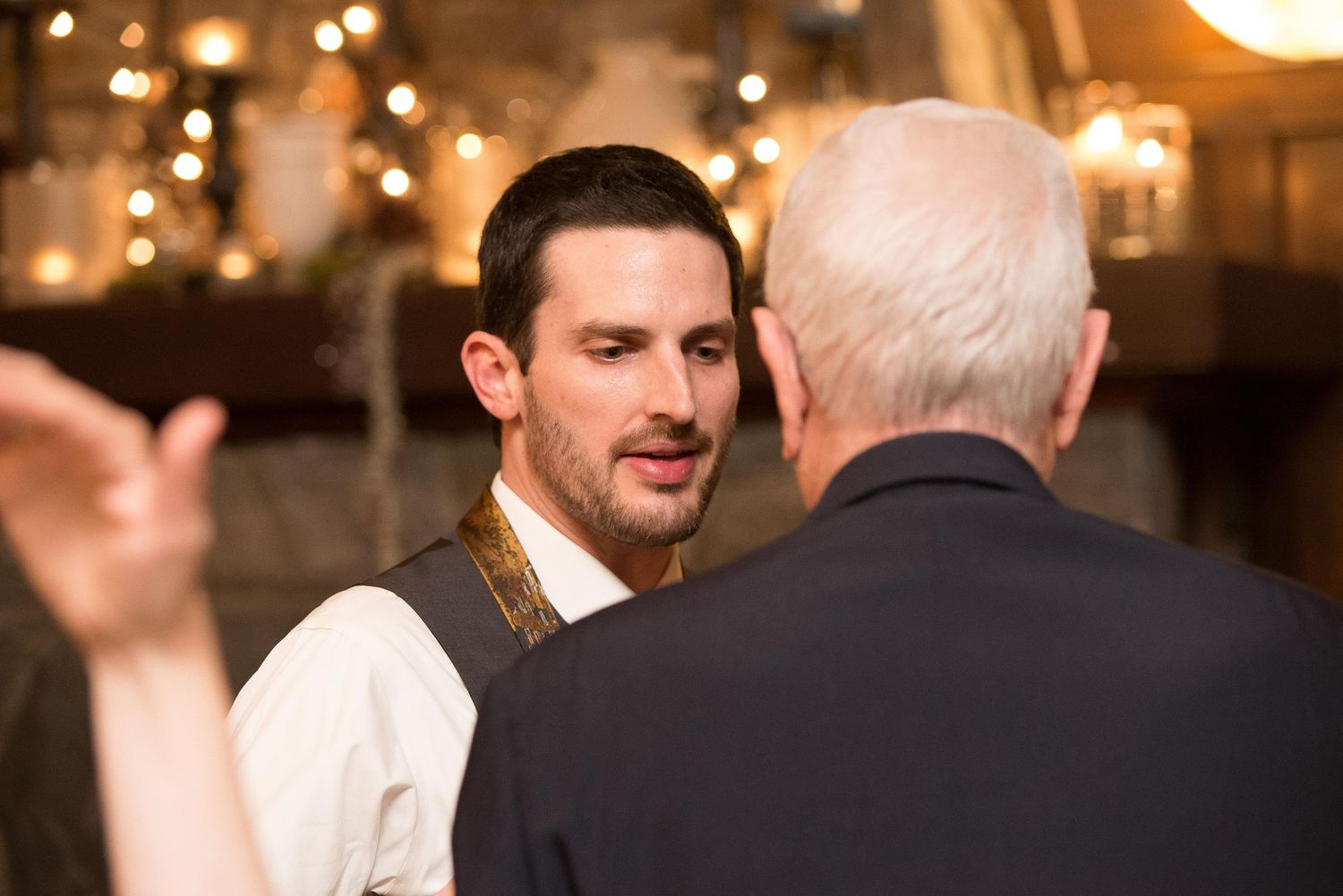 Two men in formal attire engaged in conversation at an elegant indoor event with warm string lights in background.