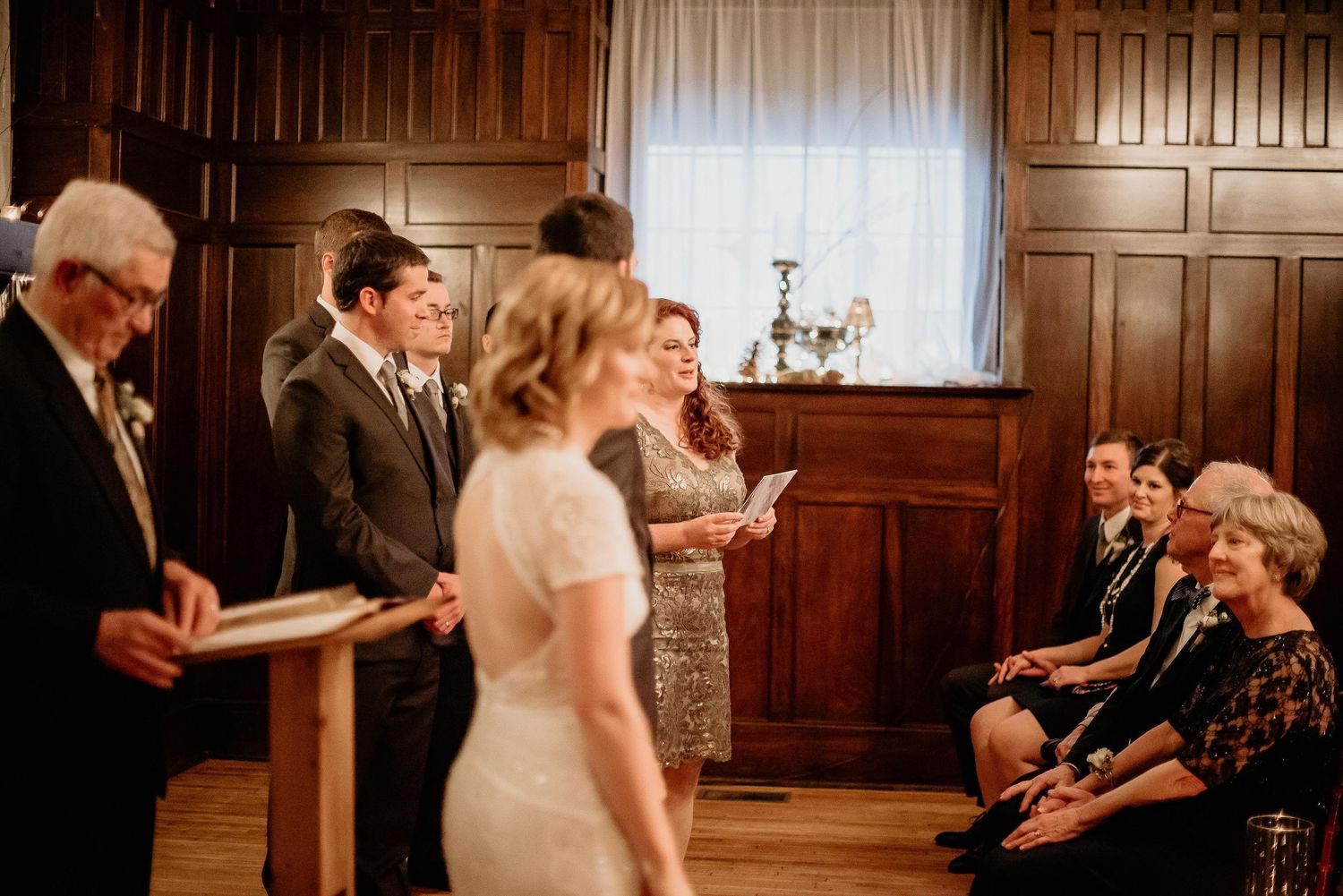Elegant wedding ceremony in wood-paneled room with bride, groom, and officiant exchanging vows before seated guests.