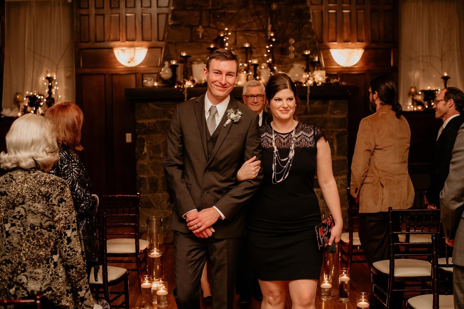 Couple smiling together at an elegant indoor wedding ceremony with candlelight and a stone fireplace backdrop.