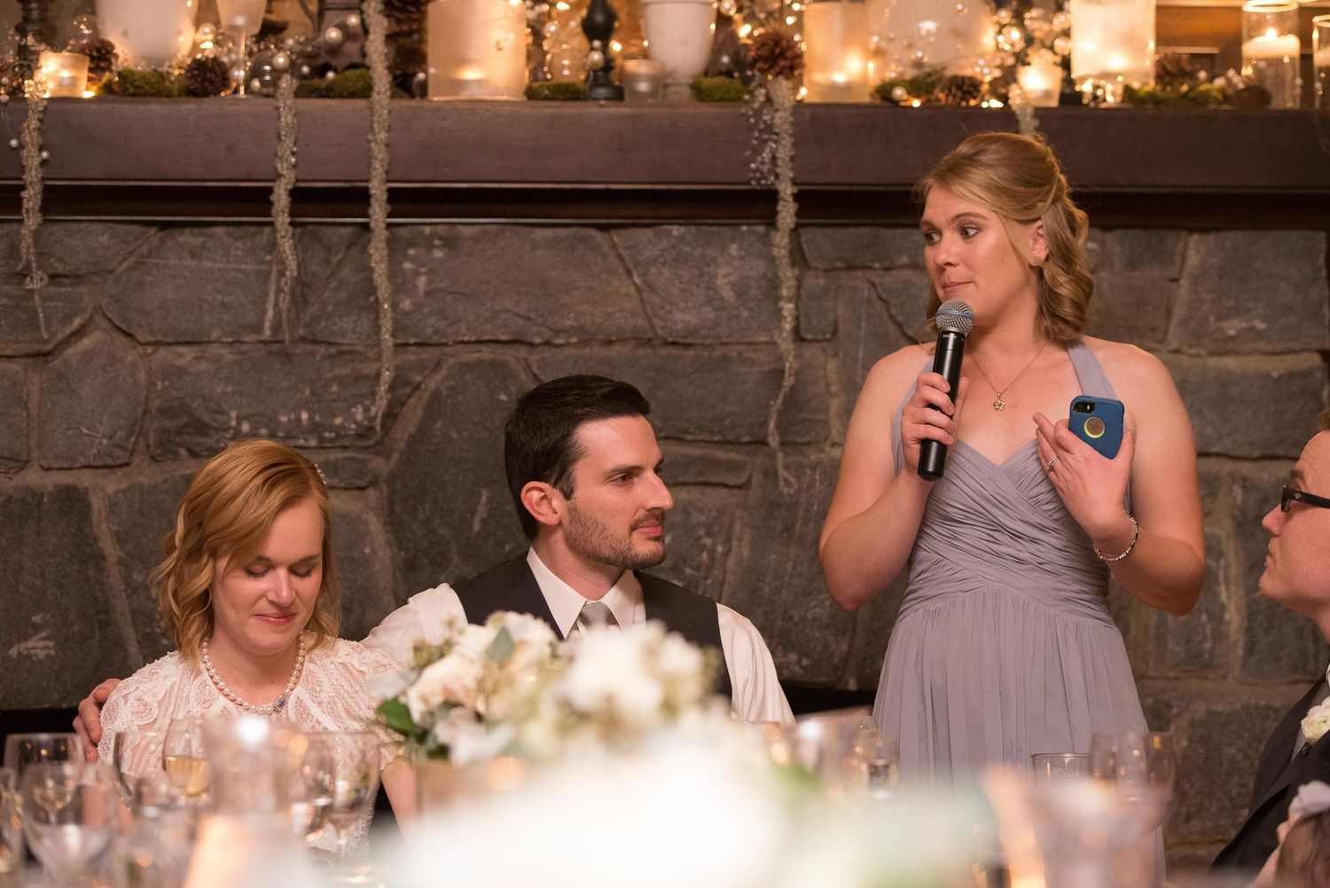 Bridesmaid giving a speech at a wedding reception with microphone, seated guests at decorated table.