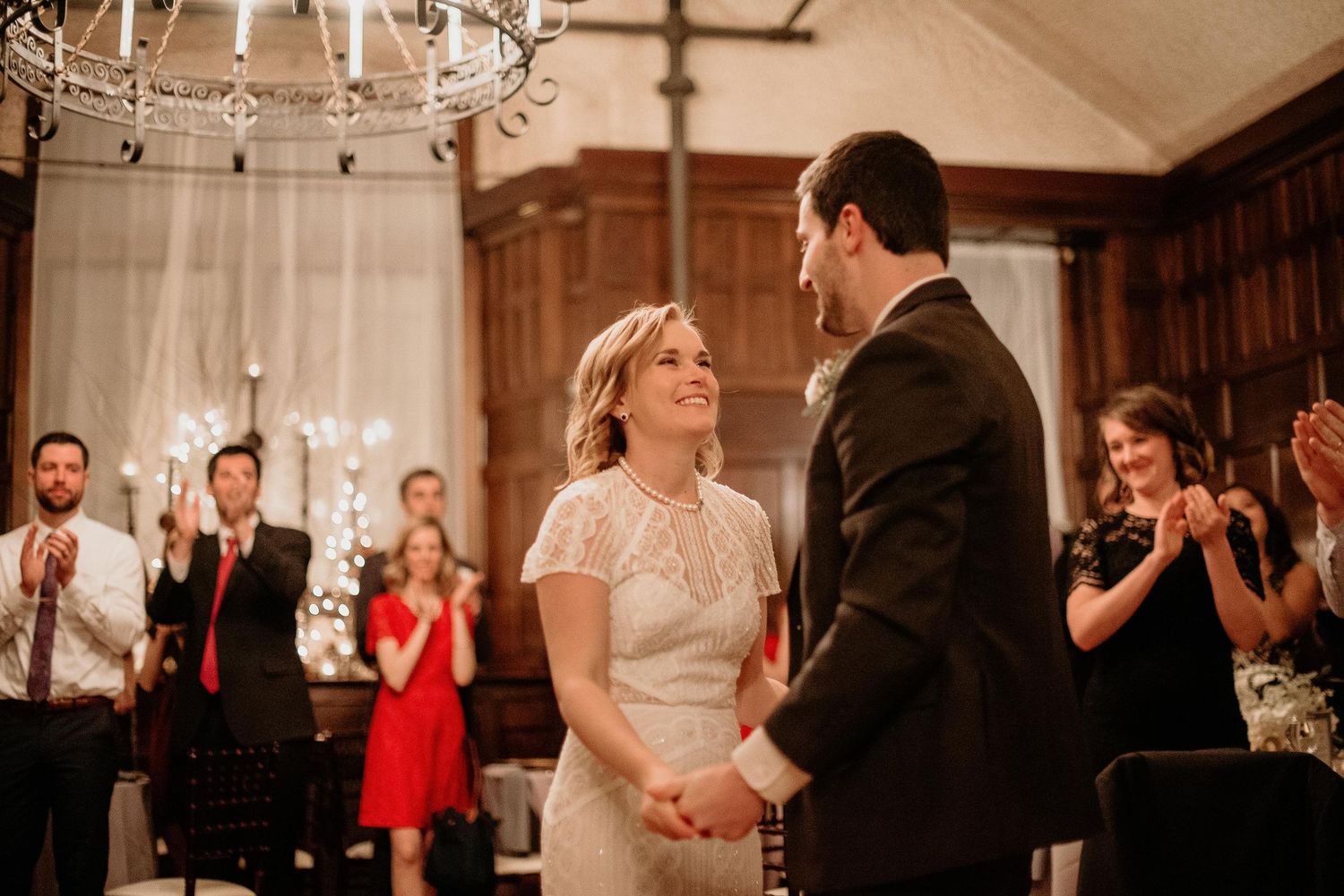 Bride and groom share first dance while wedding guests clap and celebrate in elegant wood-paneled reception hall.