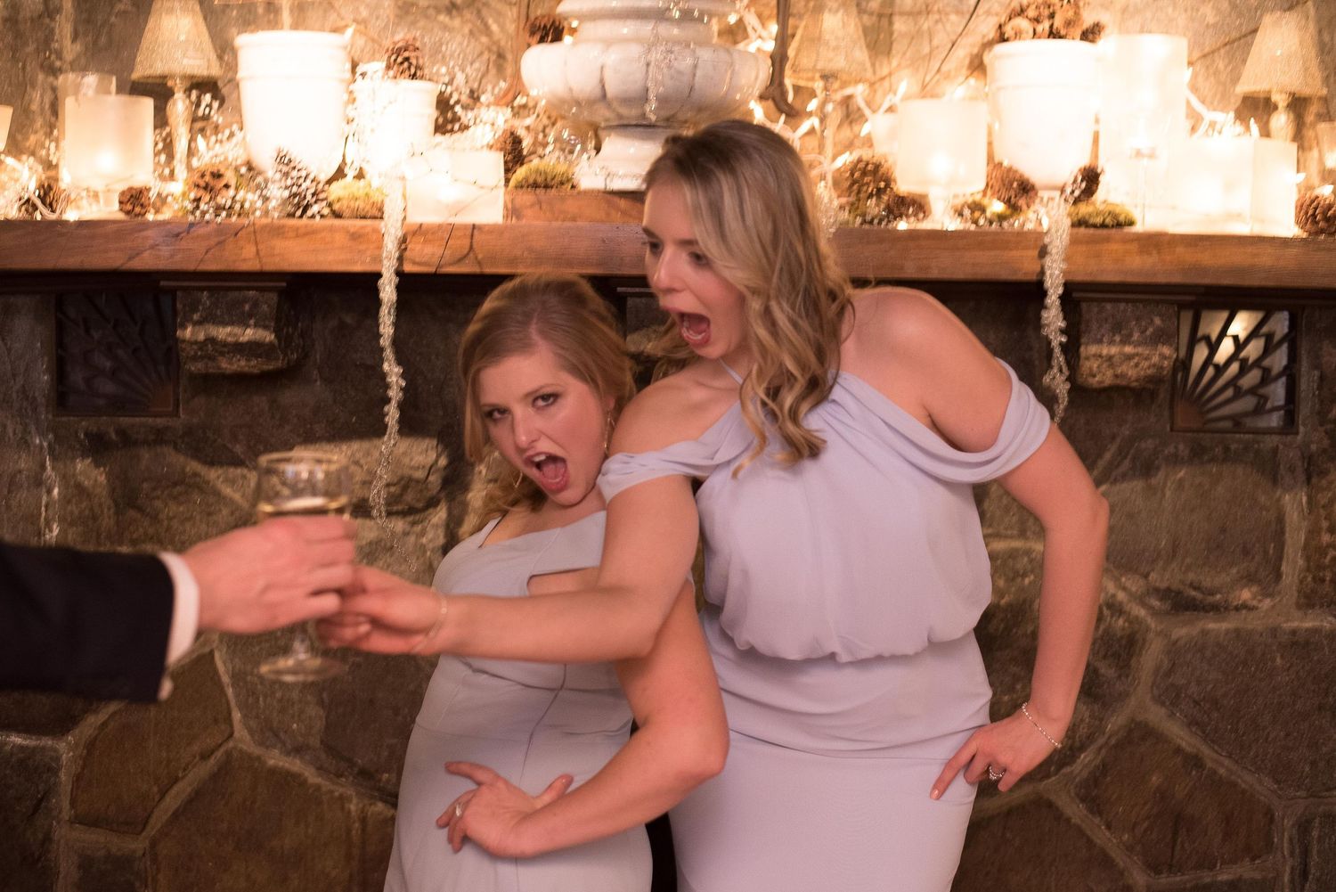 Two bridesmaids in silver dresses share a funny reaction while holding champagne at a candlelit wedding reception.