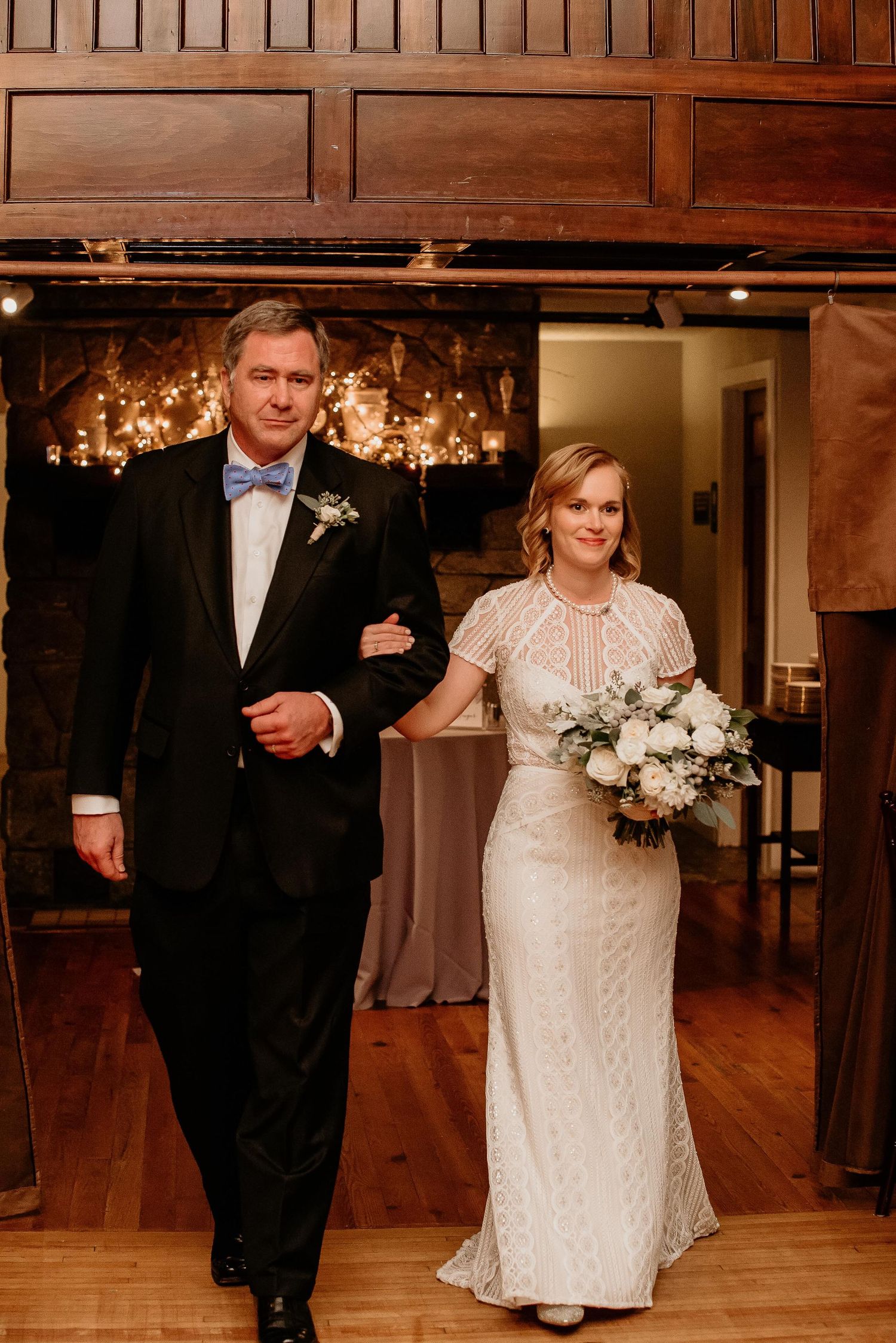 Bride in lace gown holding white bouquet walks arm-in-arm with man in tuxedo and blue bow tie at wedding reception.