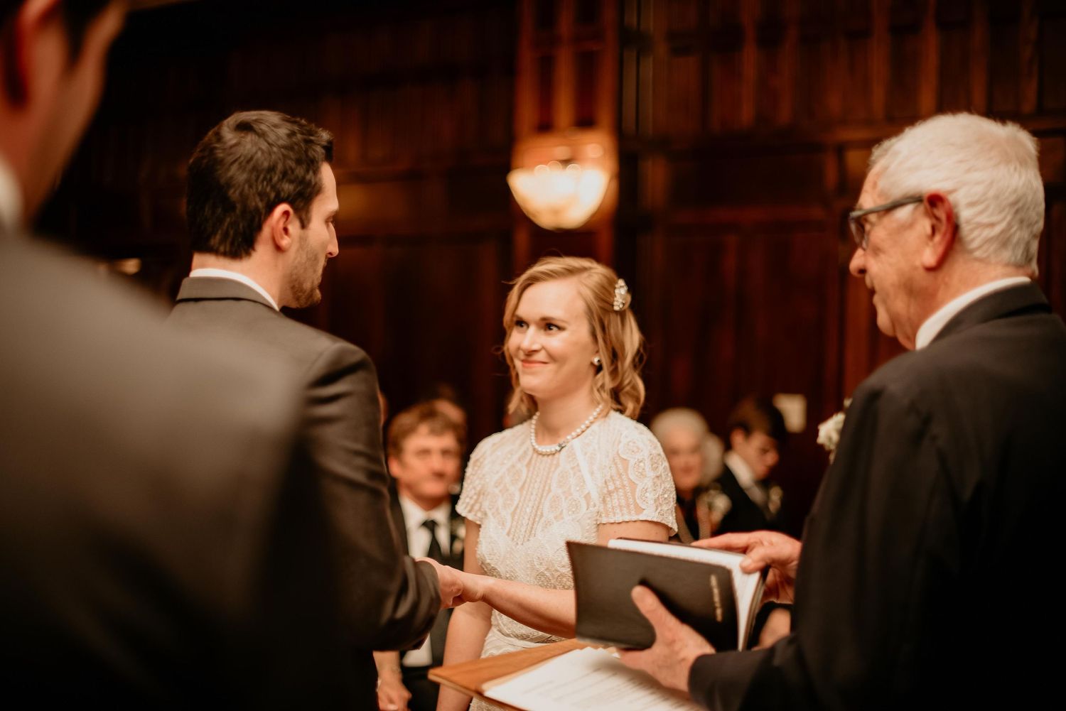 Bride smiling joyfully at groom during indoor wedding ceremony with officiant holding a book.