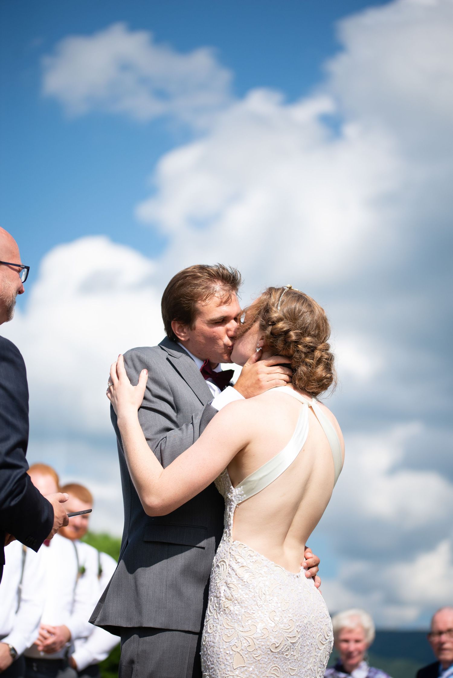Newlyweds share first kiss outdoors under bright blue sky with fluffy white clouds.