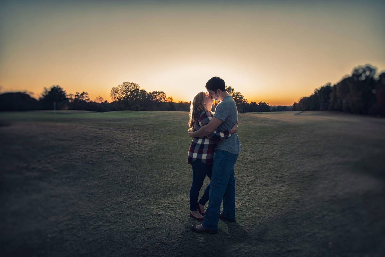 A couple embraces silhouetted against a stunning golden sunset in an open field during an engagement session.