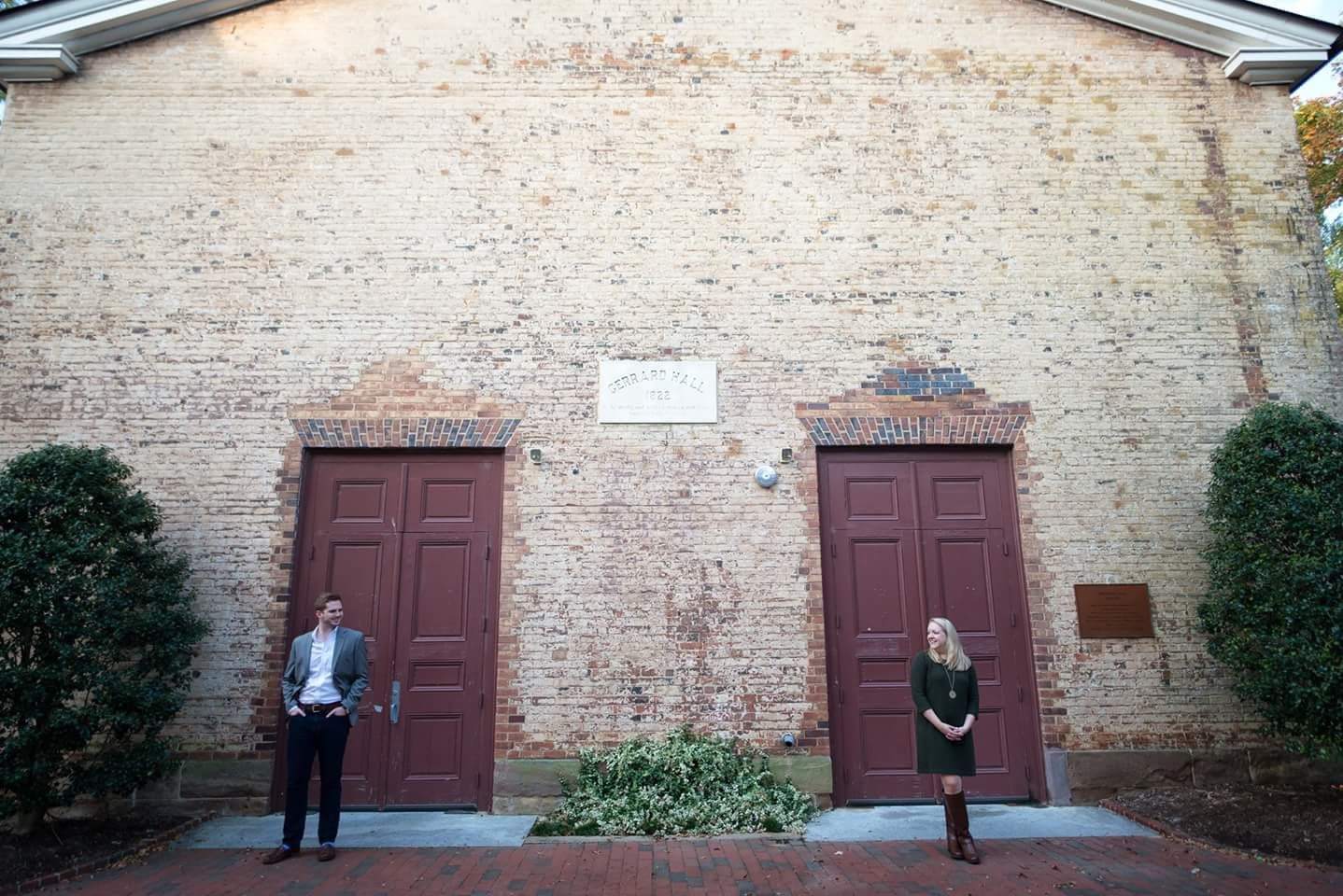 Engaged couple poses apart before historic brick building with red doors.