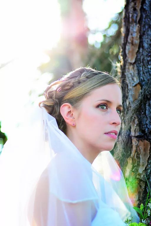 Beautiful bride with braided updo and veil posing beside a tree in soft sunlight.