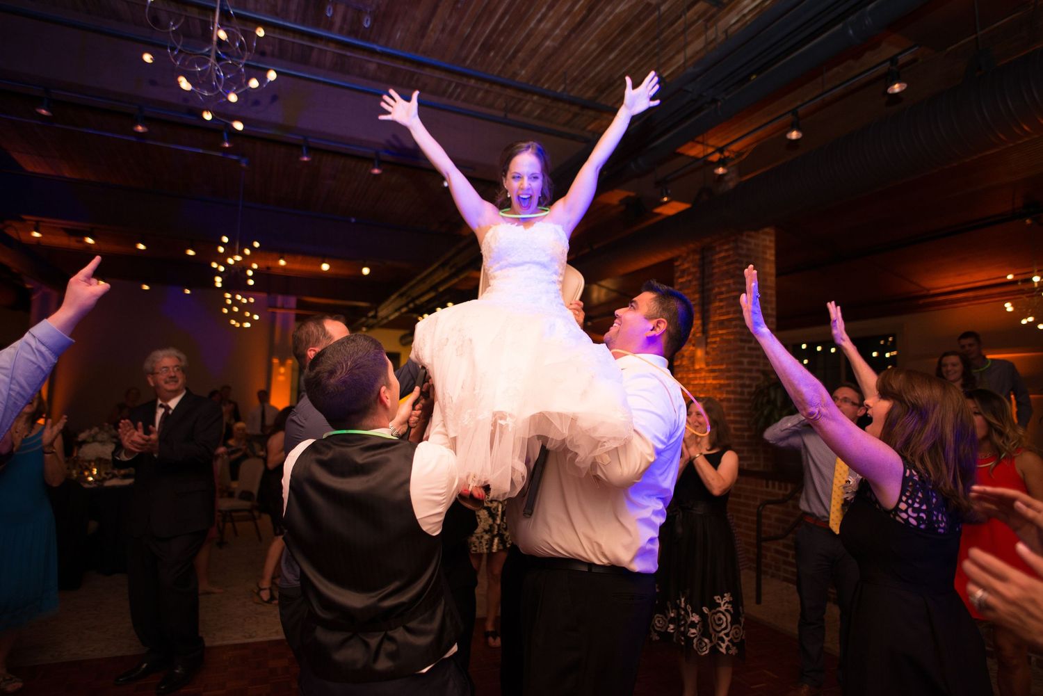 Bride lifted by guests on wedding dance floor, arms raised joyfully.
