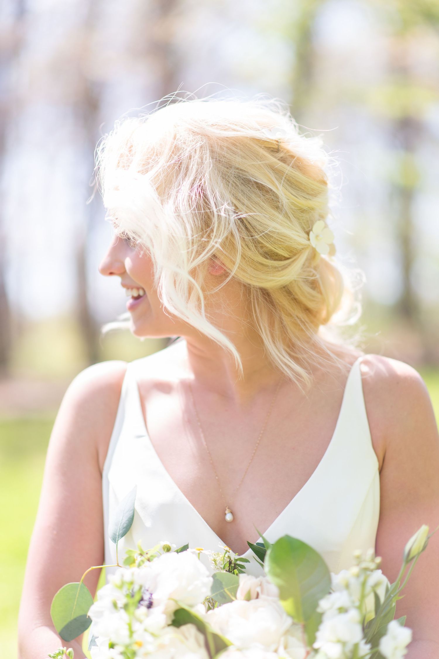 Smiling blonde bride with updo hairstyle holds white floral bouquet in sunlit garden.