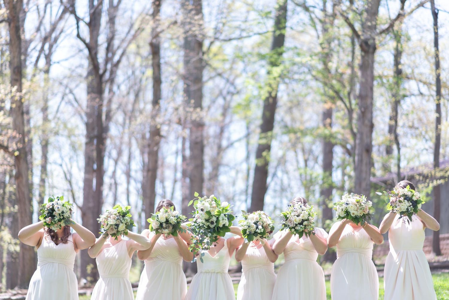 Row of bridesmaids in white dresses holding lush green and white bouquets overhead.