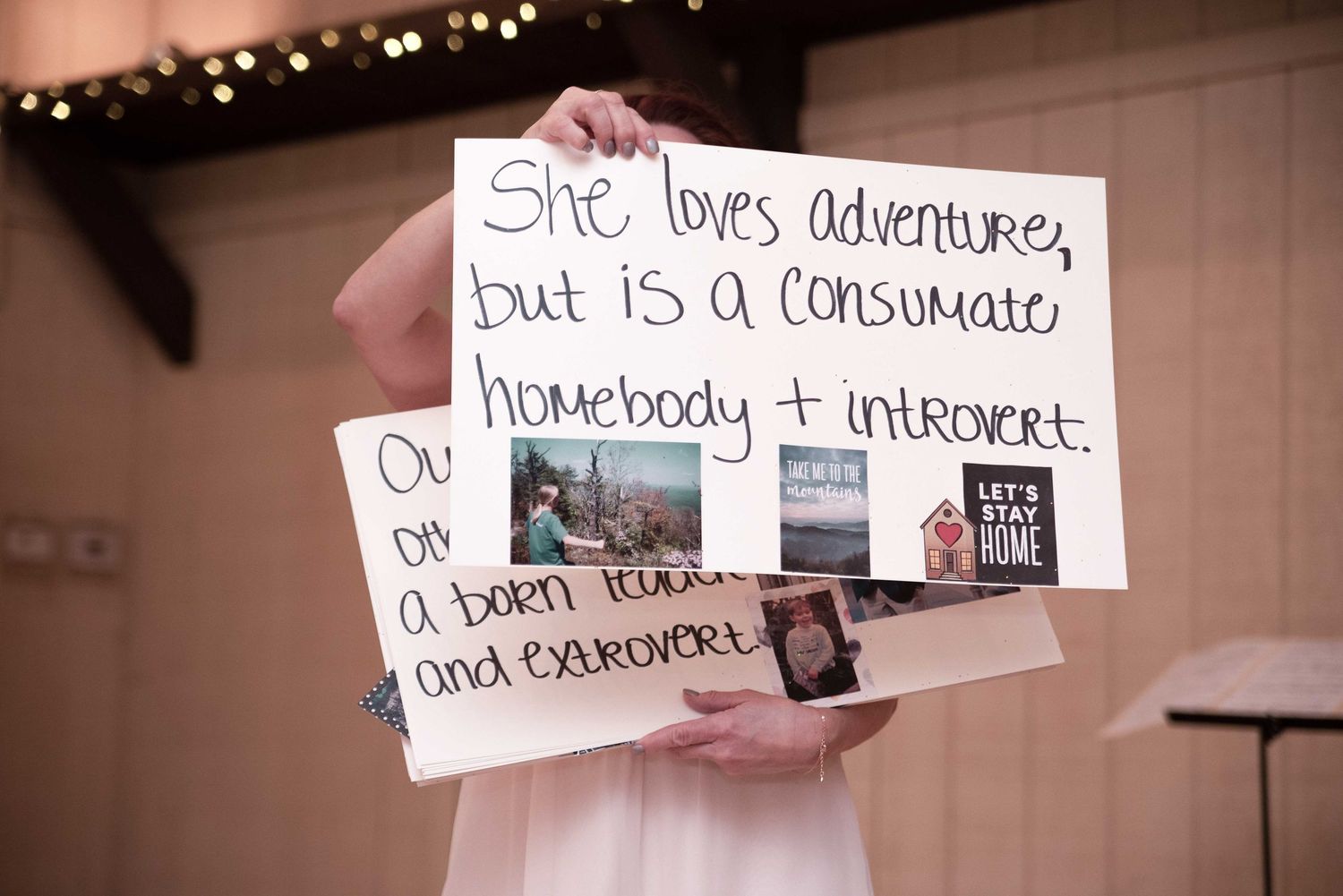 Person holding a sign reading 'She loves adventure, but is a consummate homebody + introvert' at a wedding.