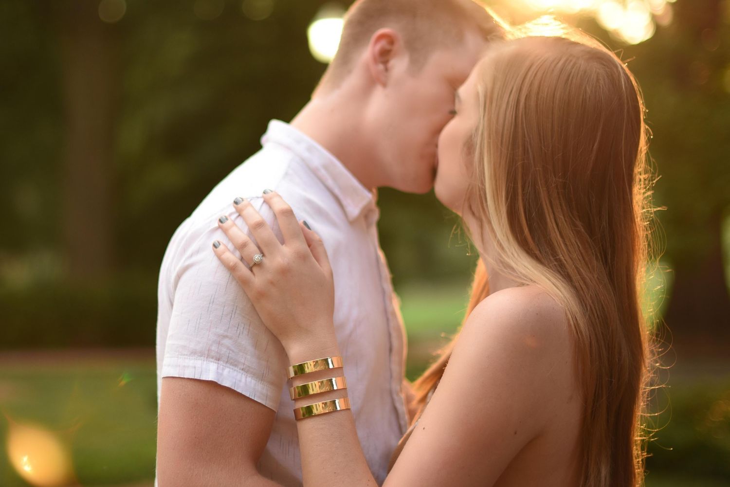 Romantic couple sharing a tender kiss outdoors in golden sunset light, woman wearing gold rings and bracelet.