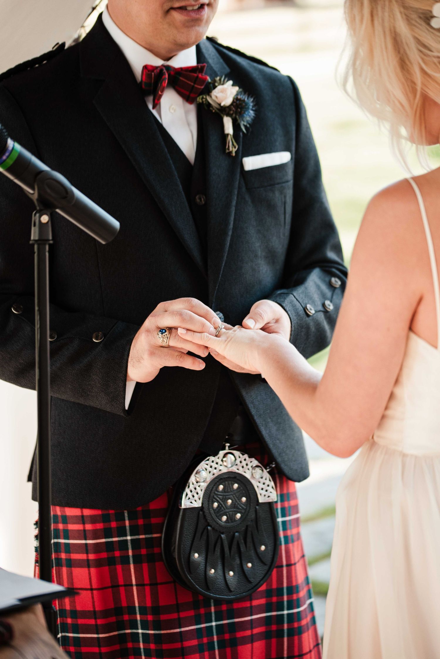 Groom in Scottish Highland dress with red tartan kilt and sporran exchanges wedding rings with bride during ceremony.