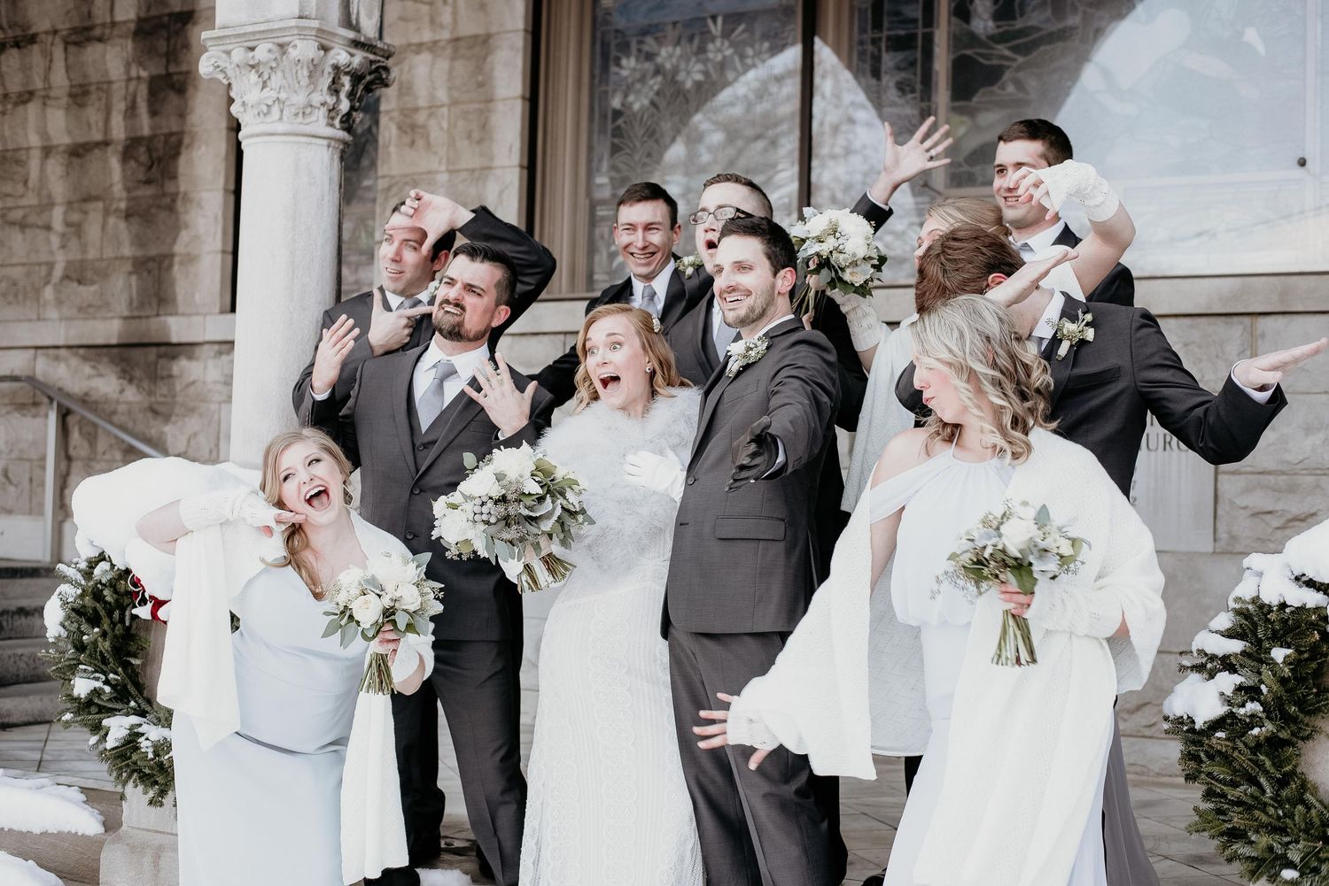 Wedding party celebrating outdoors in winter, bridesmaids in white dresses holding bouquets, groomsmen in gray suits, joyful poses.