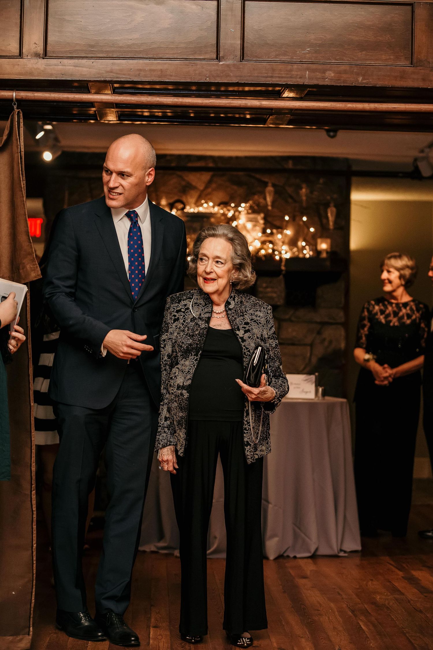 Elegant man in navy suit escorts older woman in black outfit at formal indoor event near stone fireplace.