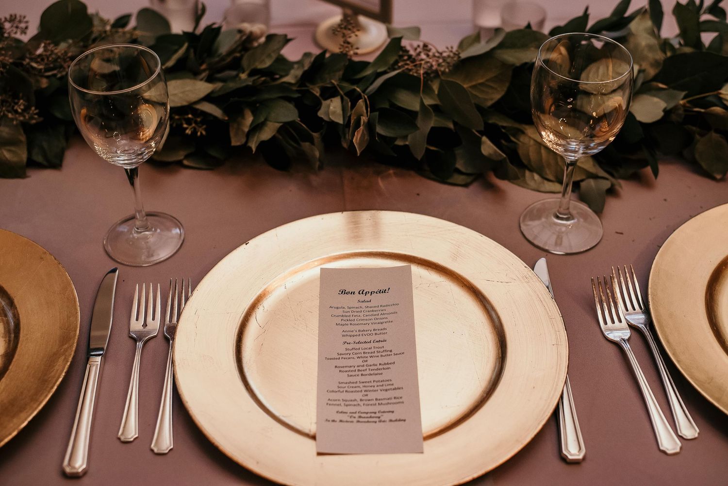 Elegant gold charger plates with silverware, crystal wine glasses, and a menu card on a mauve tablecloth with greenery.