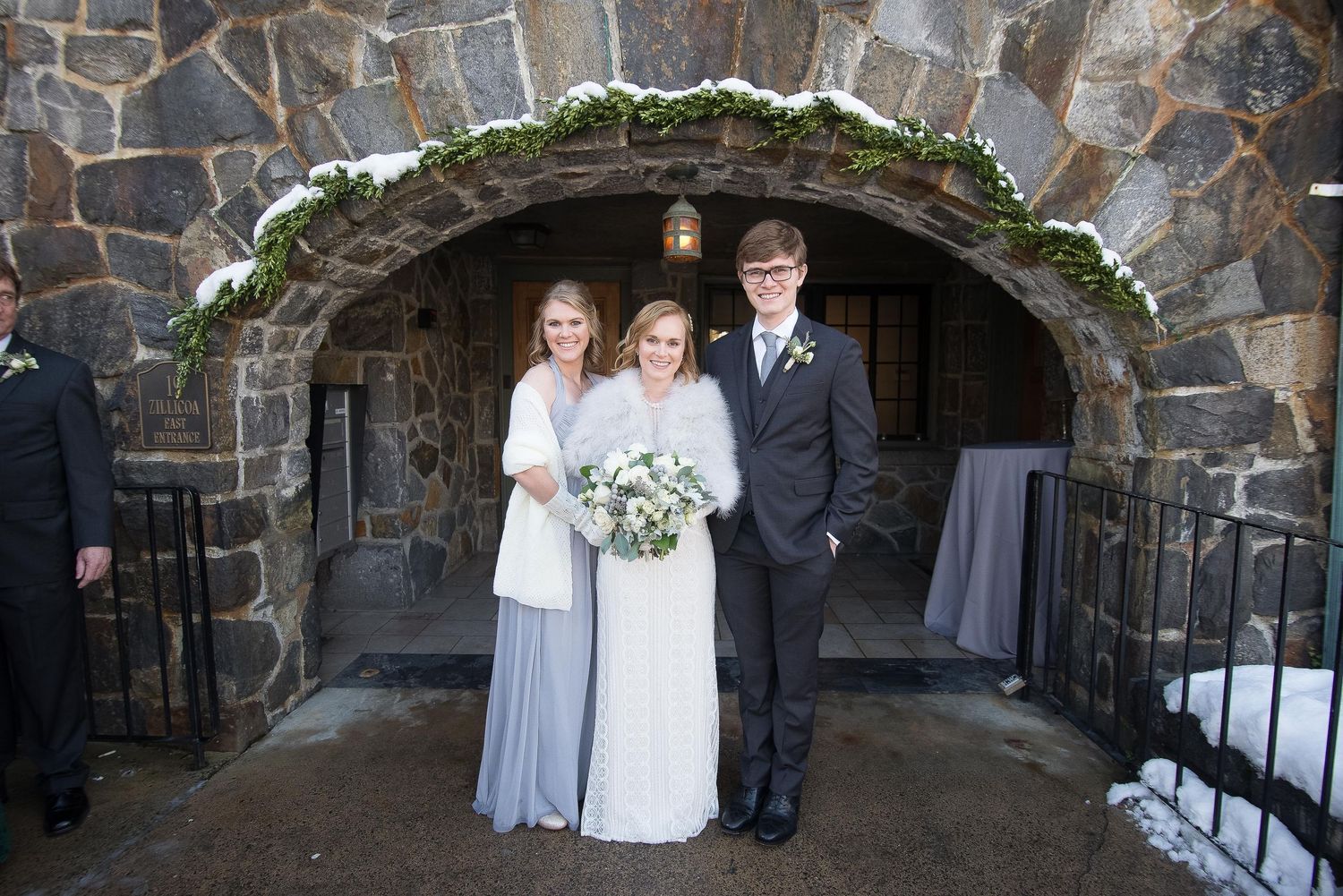 Bride in white gown with two guests under stone arch decorated with greenery at winter wedding venue.