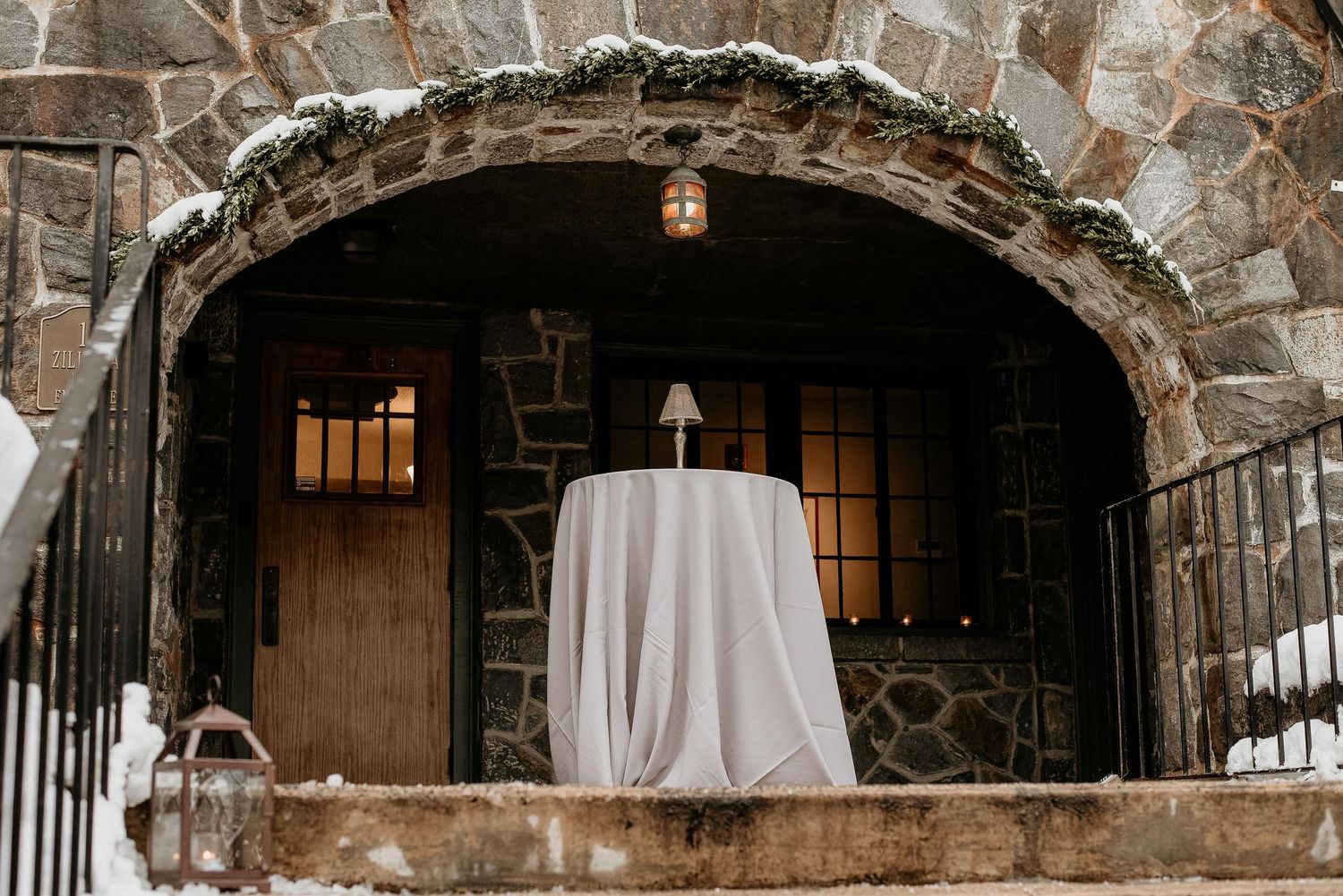 Elegant winter wedding venue entrance with stone arch, draped table, and lanterns dusted with snow.