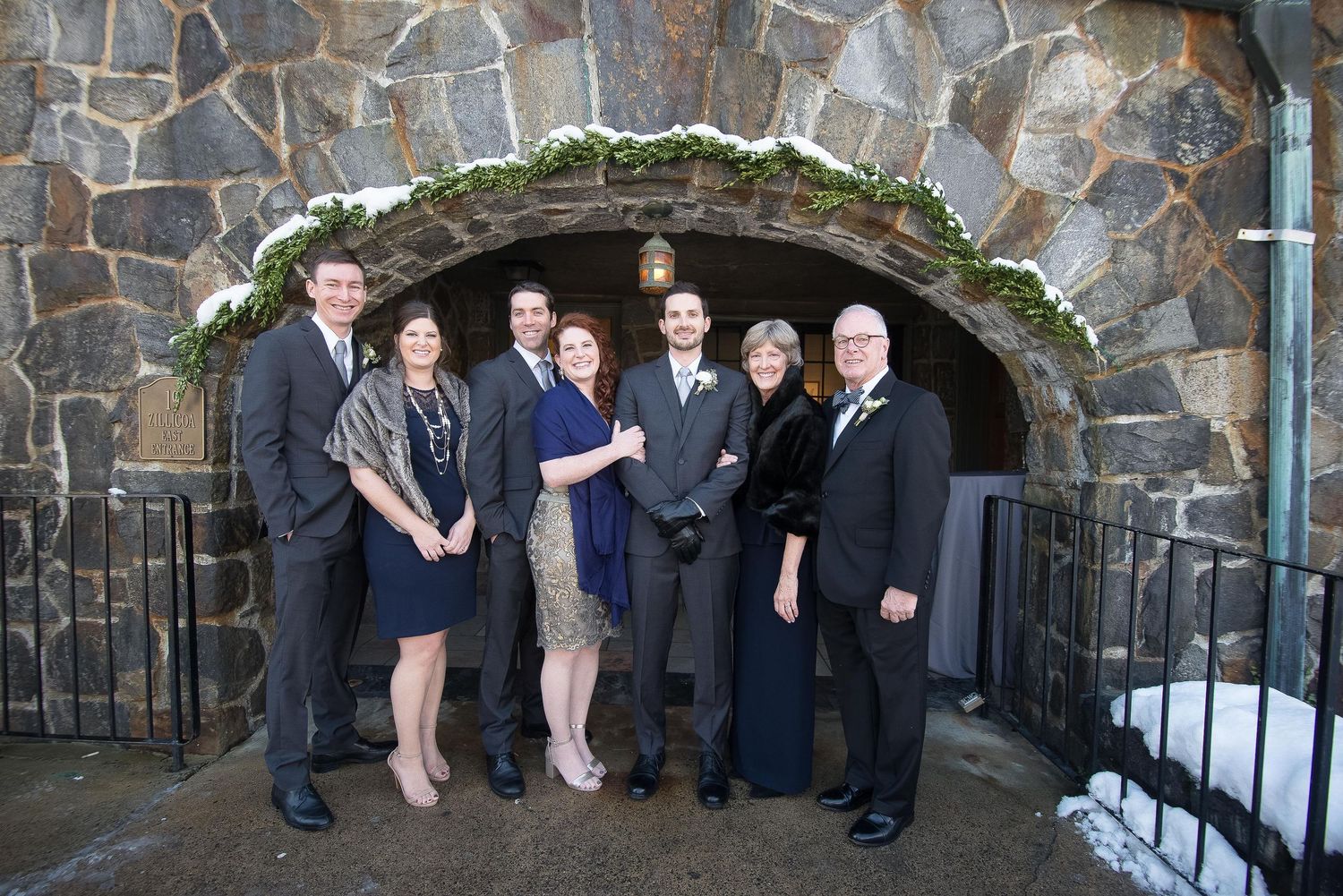 Group of seven formally dressed people posing together under a stone arch at an outdoor winter wedding venue.