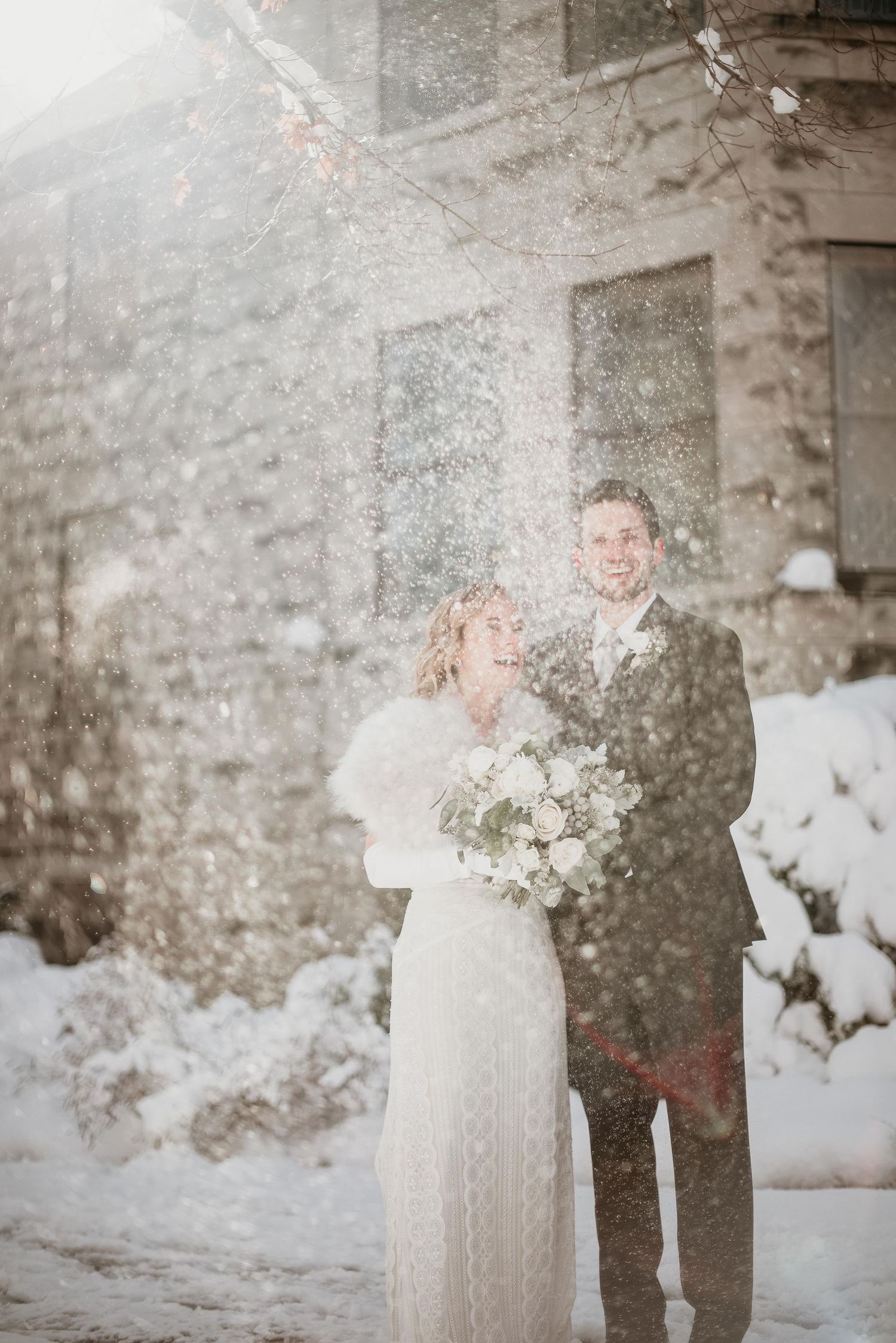 Bride and groom smiling together in a winter snowstorm, bride holding white bouquet outside stone building.