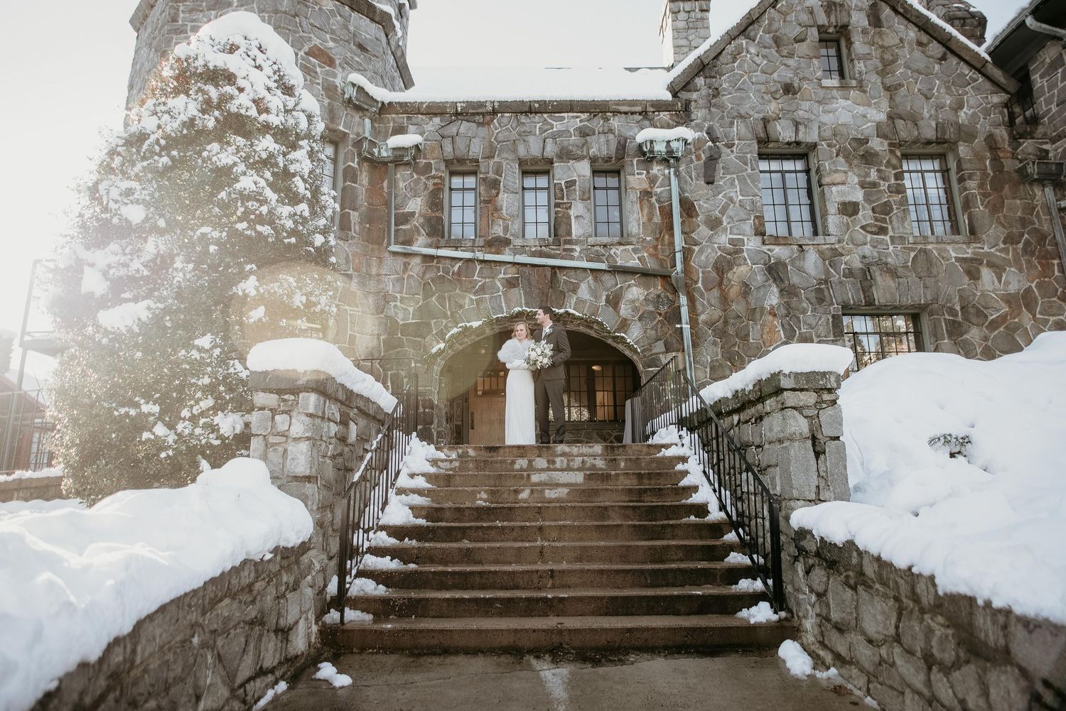 Bride and groom kissing on snowy stone castle steps during winter wedding ceremony.