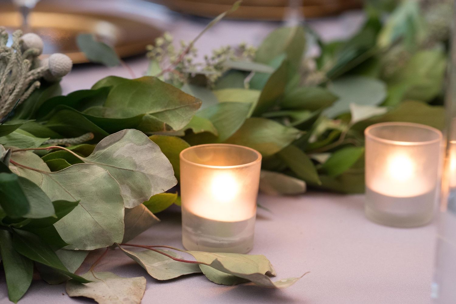 Glowing votive candles nestled among fresh eucalyptus leaves on an elegant wedding reception table.