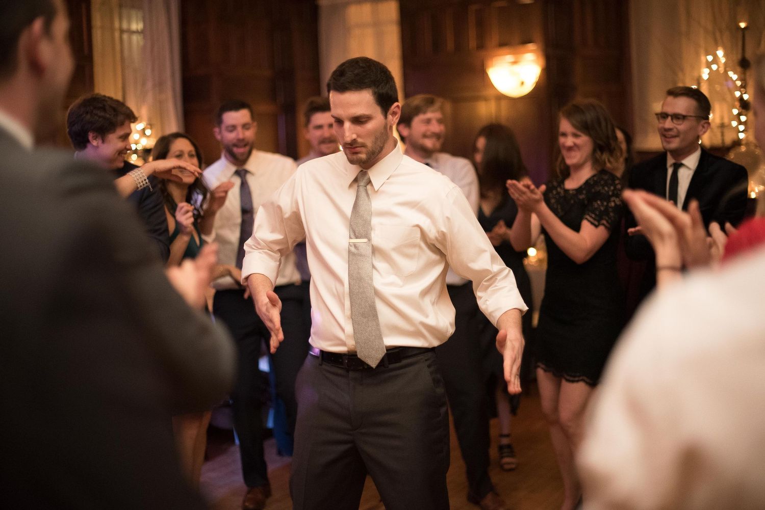Groom dancing energetically at wedding reception while guests clap and celebrate in elegant venue.
