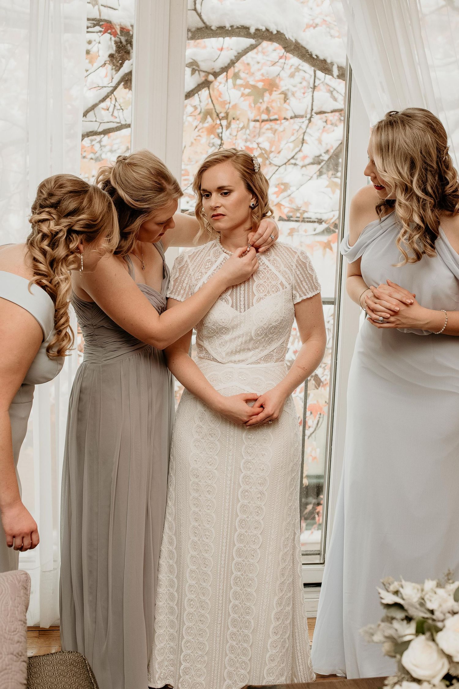 Bridesmaids in gray gowns help bride in white lace dress prepare near a window with autumn foliage.